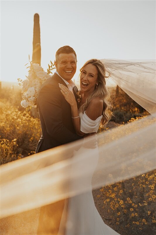 A couple stands in a field of wildflowers in Tucson, Arizona, smiling at the camera as the setting sun illuminates their surroundings.