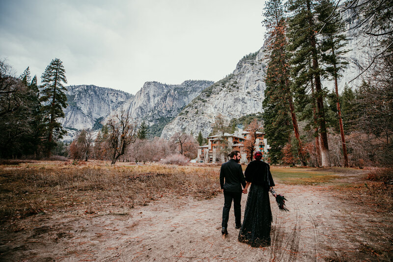A couple dressed in elegant black wedding attire walks hand in hand toward the historic Ahwahnee Hotel, surrounded by towering cliffs and trees in Yosemite National Park.