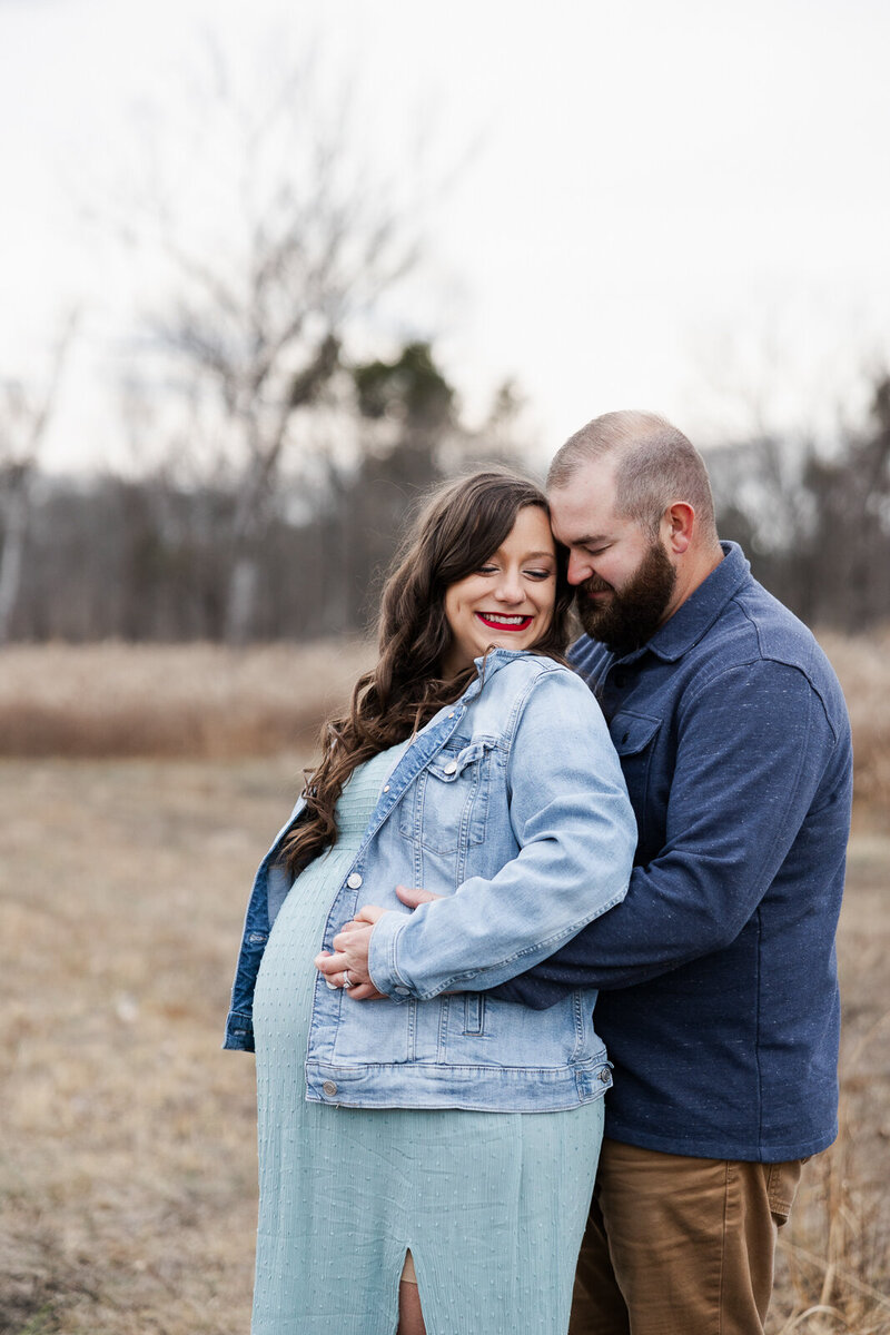 Portrait of a couple who are both wearing blue jean tops with their hands on mom's belly looking at each other smiling. 