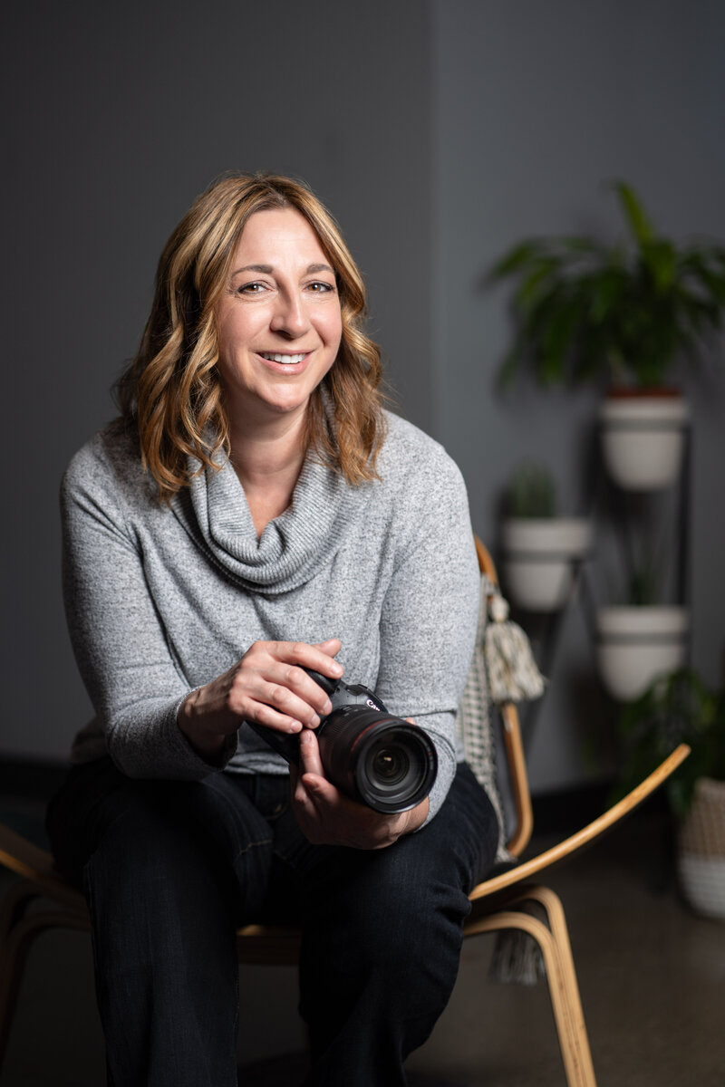 A female photogapher is seated on a wood chair with a shadowed gray wall and plants behind her.  She is wearing a light gray sweater and is holding a Canon camera