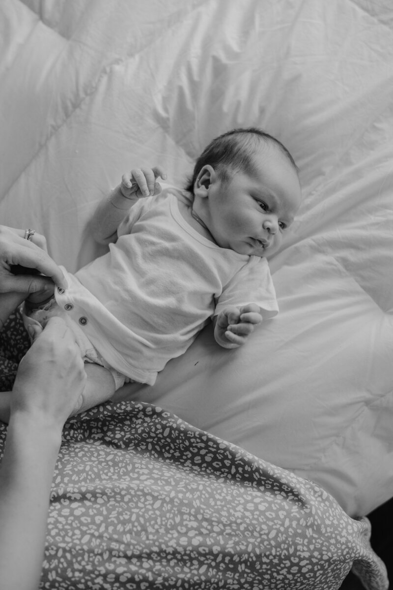 Black and white photo of mom changing baby 's onesie while baby is laying on the bed in a studio in Annapolis Maryland photographed by Bethany Simms Photography