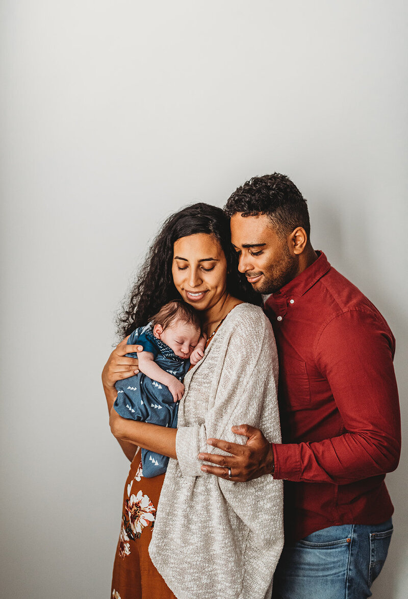 in studio session with mother and father in coordinating orange outfits while the mother holds their baby in a blue swaddle while the father stands behind the mother and holds her while looking over her shoulder at the baby captured by Family photographers Maryland