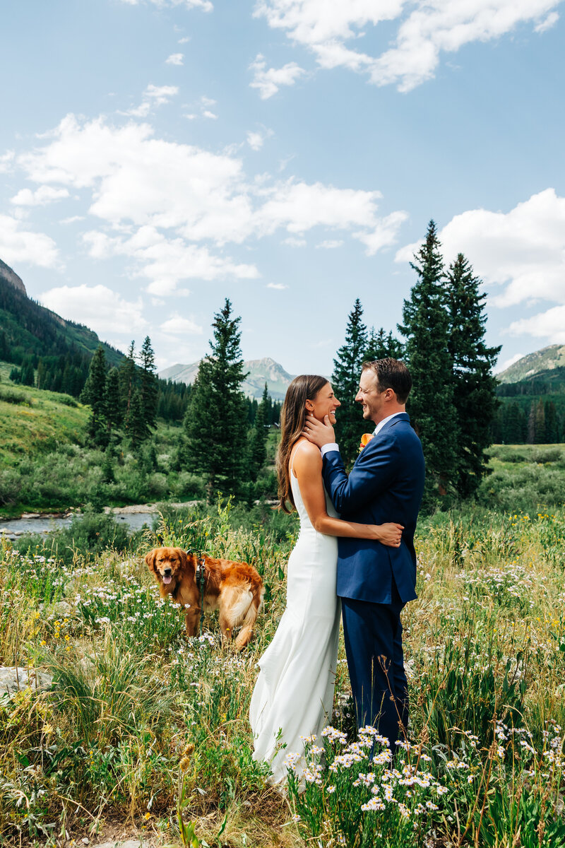 bride and groom are looking at the camera holding their dog. The dog has a flower collar and there are mountains behind them with a little lake. It is very green everywhere