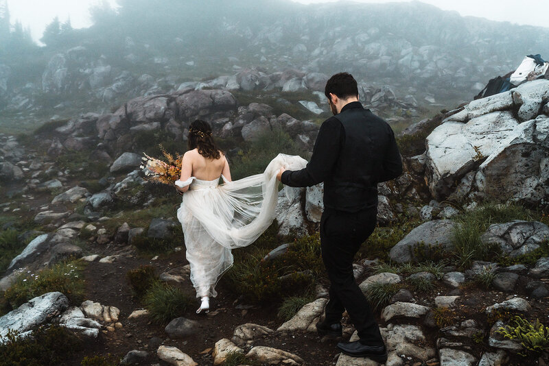 a couple in their wedding attire explore a rocky terrain. Our bride leads the way as her groom follows holding the train of her wedding dress. 