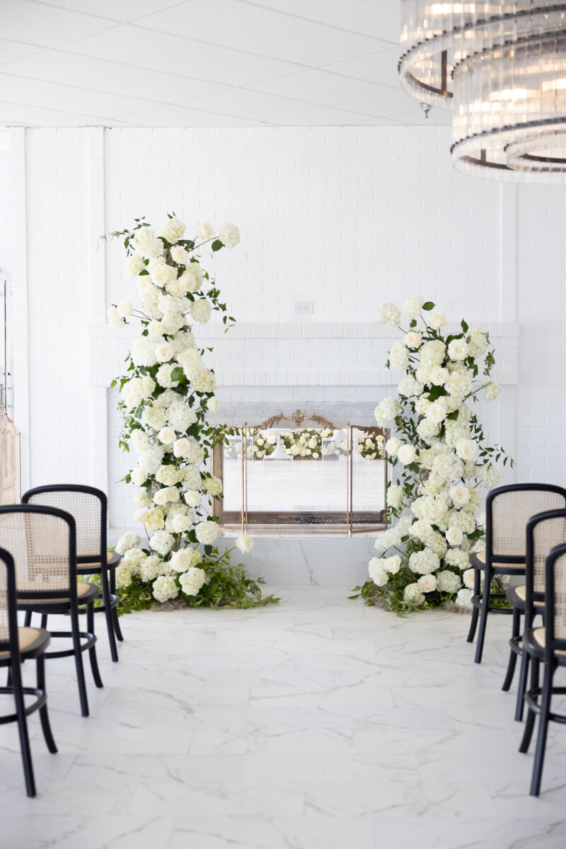 White floral pillars framing a fireplace with black cane-back chairs in a bright indoor venue.