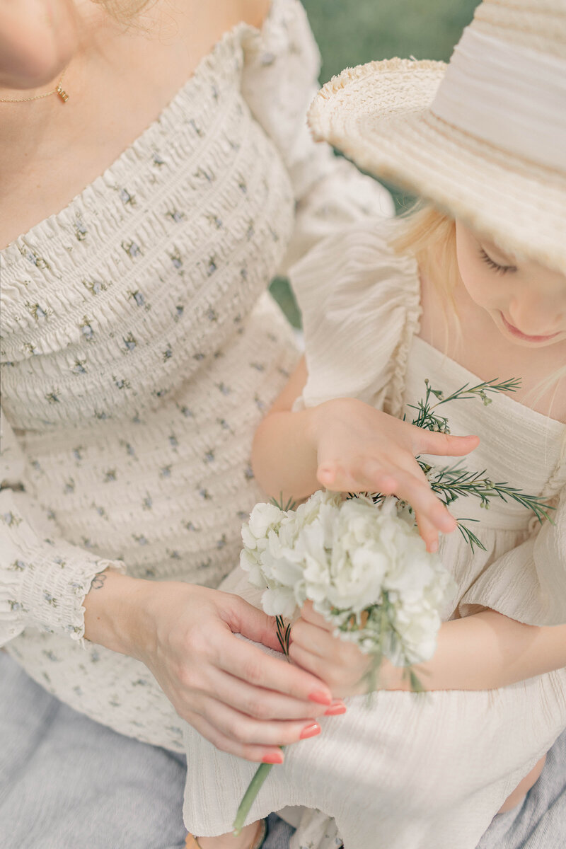 Close-up of a little girl in a straw hat sitting on her mother’s lap while delicately holding white flowers during a Kansas City mommy-and-me session.