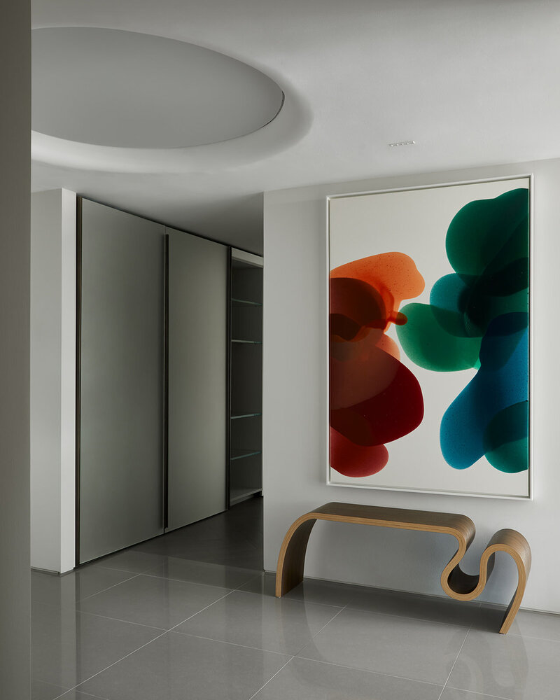 A minimalistic foyer area featuring polished grey stone tile flooring, an artistically curved wood bench, a large round lighting fixture, and colourful abstract wall art