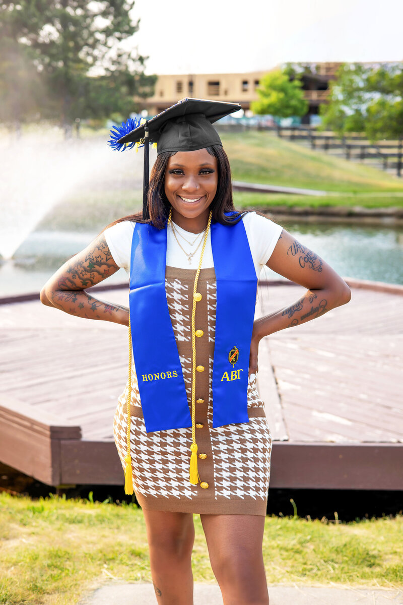 College grad wearing black graduation cap, blue honors sash, yellow cord, and brown and white dress posing in front of a pond