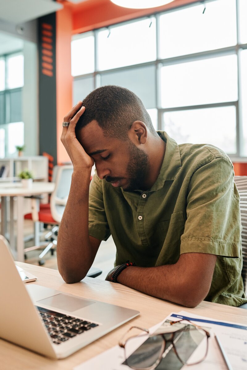 This is a solo image of a  black man sitting at a desk with his computer. He has his head resting in his hand with his eyes looking down.