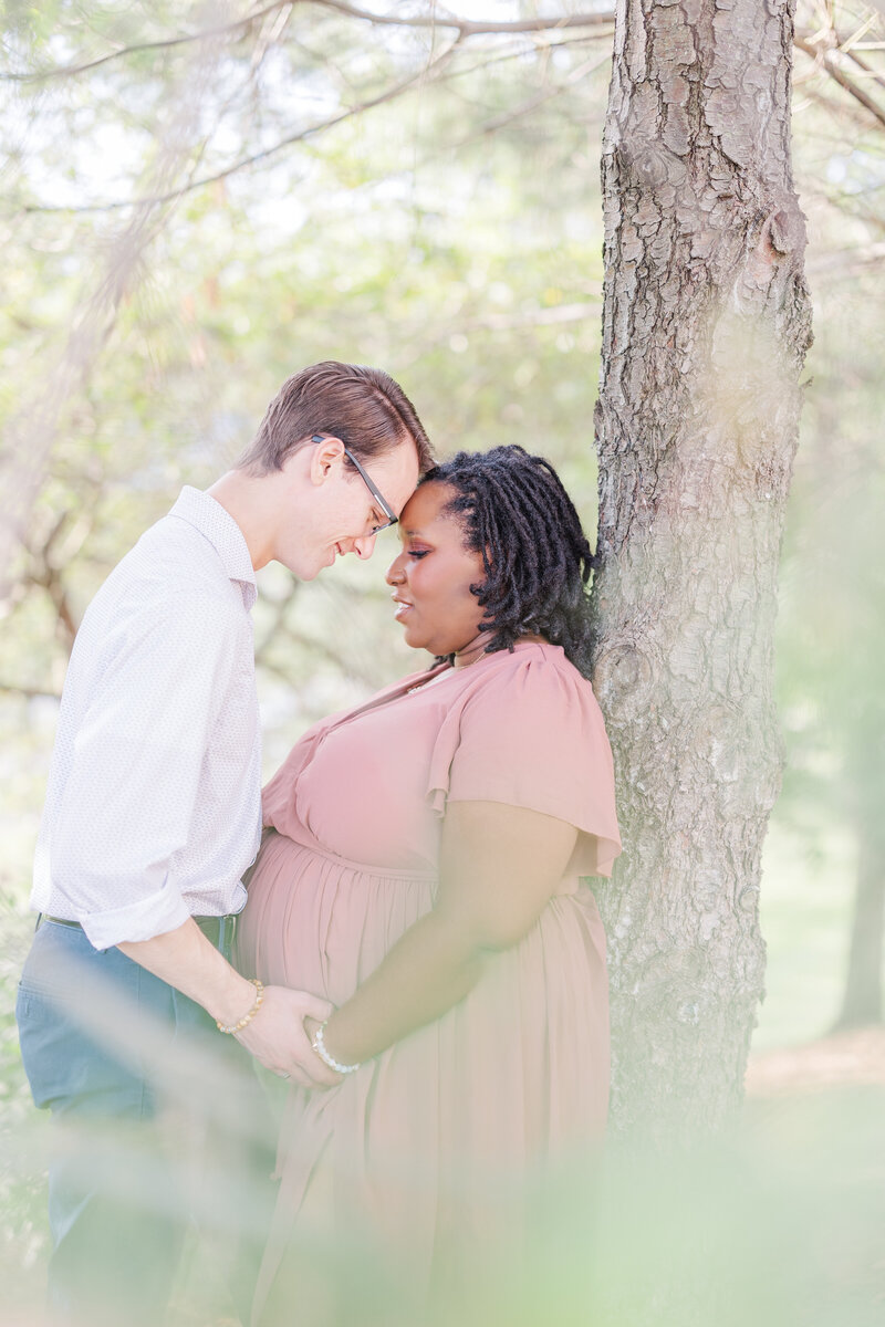 A mom to be touches foreheads with her husband while holding hands and leaning on a tree