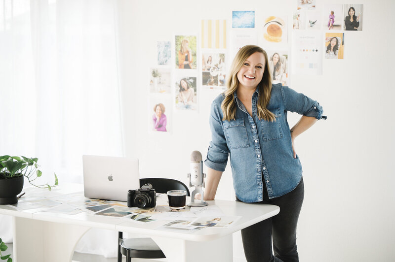 Brand photographer, Krista Marie, smiling at the camera and standing at her desk with a microphone, camera and laptop