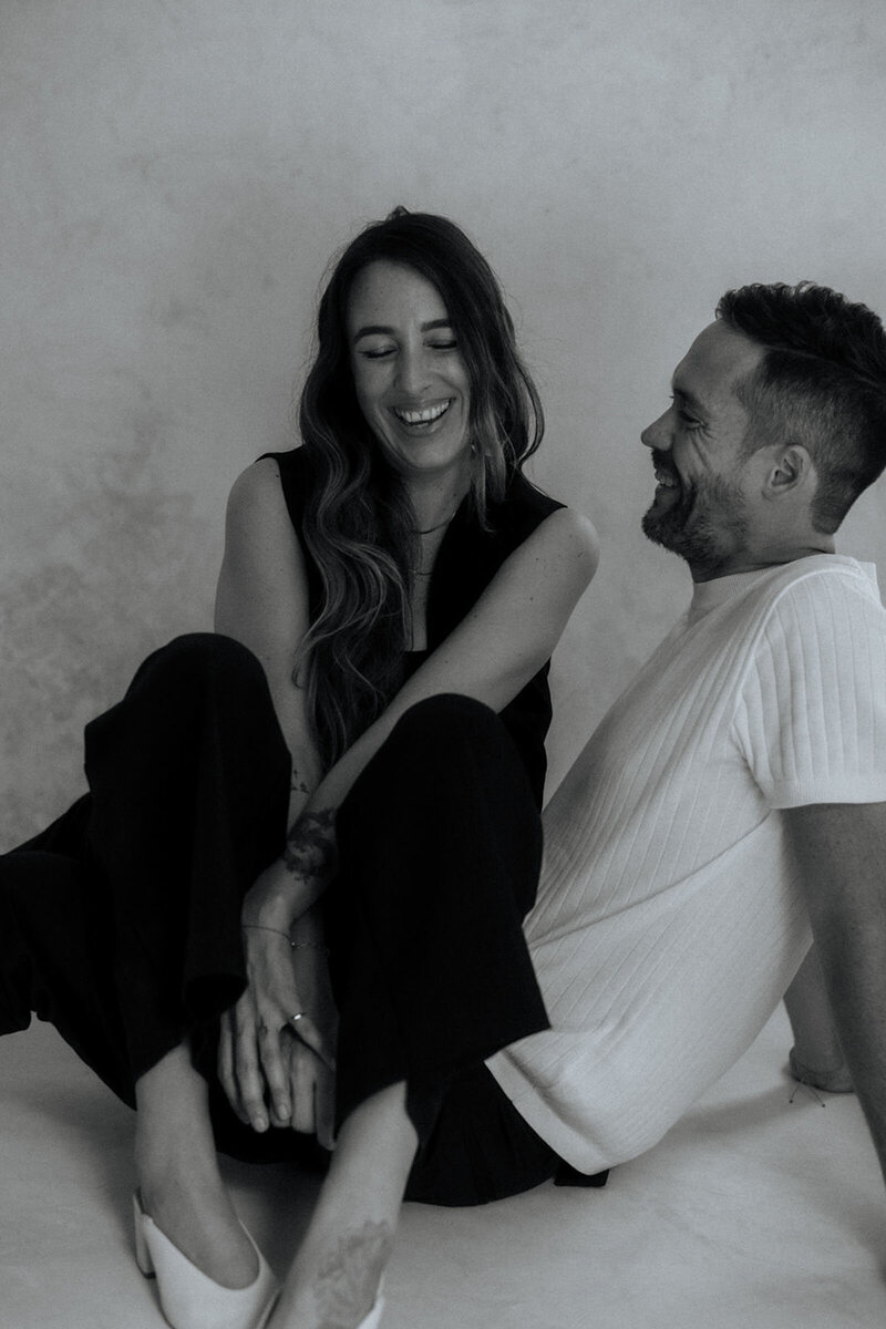 Two Cabo photographers in black and white sitting on the floor playfully draped over each other laughing