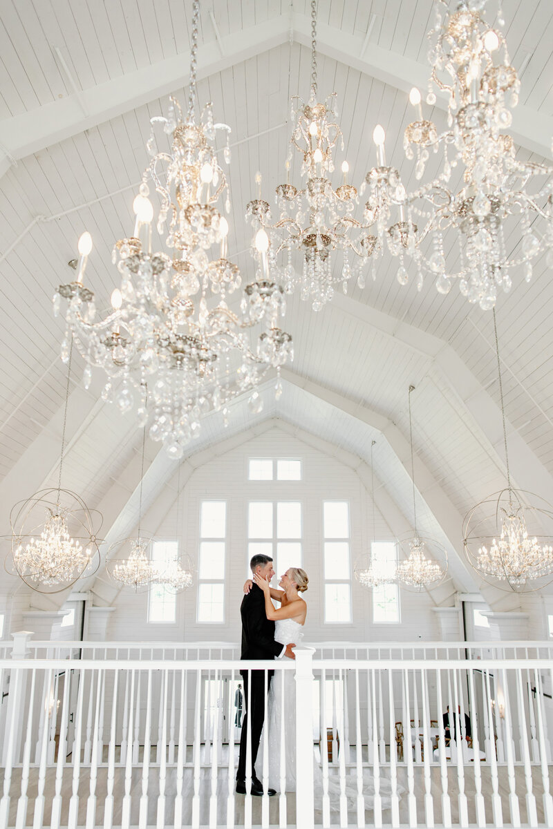 Bride and groom embrace on balcony of white barn wedding venue with chandeliers hanging