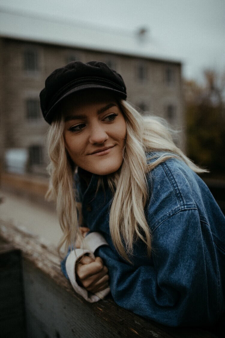 Olivia leans against a railing and smiles as she looks off camera. She's wearing a denim jacket and black cap.