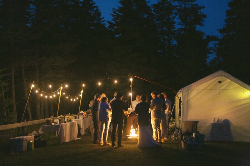 wedding guests stand around a campfire at night. Fairy lights and a glowing tent give ambient orange light.