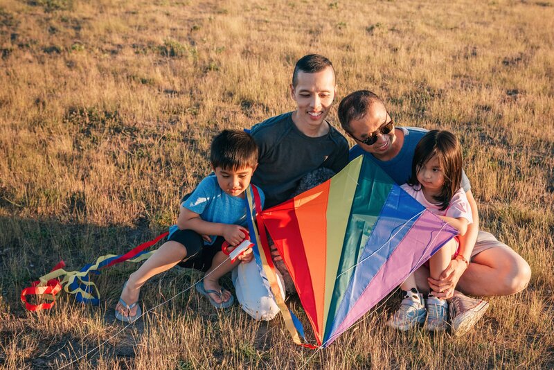 This image shows two Asian parents, both male presenting, sitting in the grass with their two young children. They are smiling and holding a rainbow-print kite.