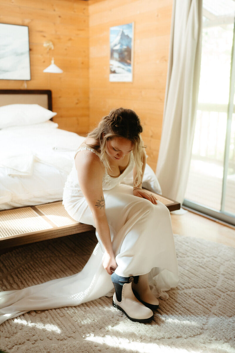 Bride getting ready for her hiking elopement, leaning down to put on her white hiking boots 