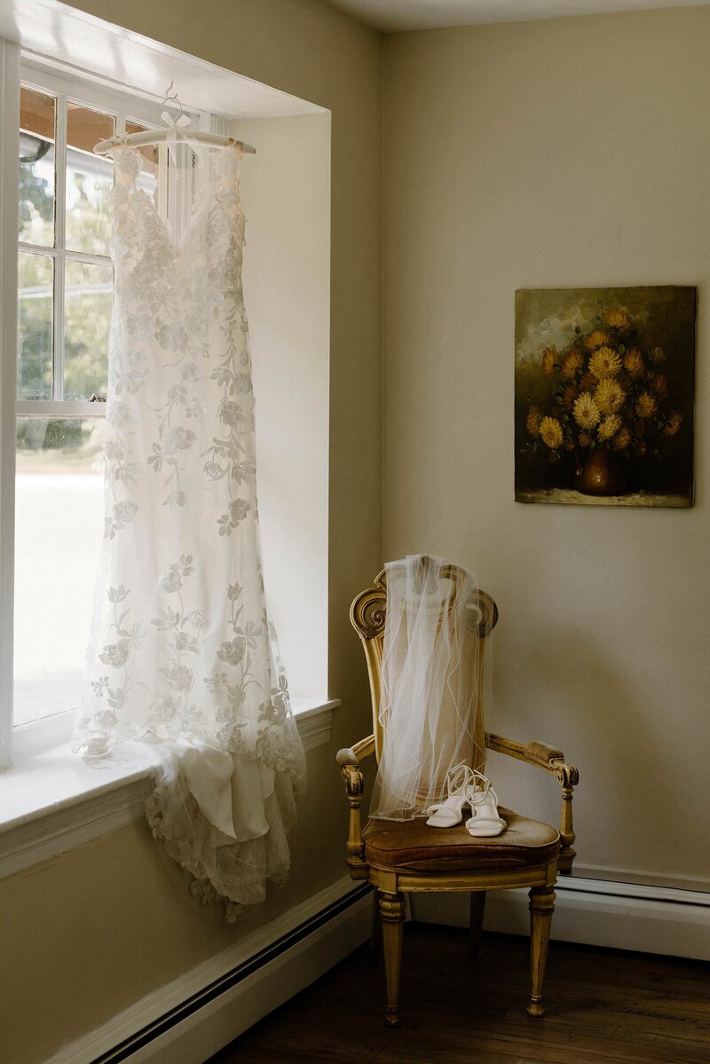 A wedding dress hanging in front of a window with a veil and shoes on a chair next to it 