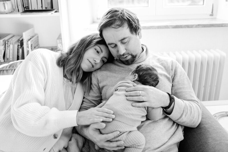 Mother rests her head against her husband's shoulder as a newborn sleeps on his chest.