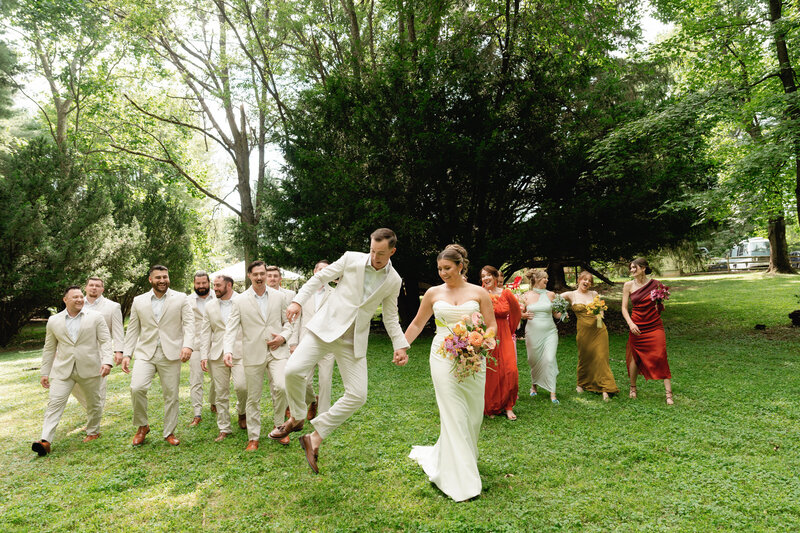 A groom clicks his heels together holding hands with his bride as their bridal party walks and laughs behind them.