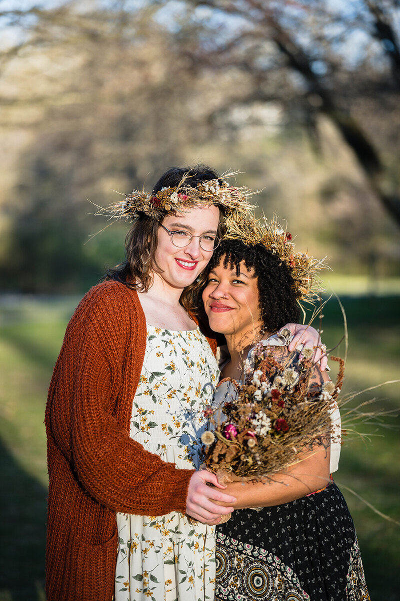 An LGBTQ+ couple on their wedding day wrap their arms around one another and smile for a photo. Each of the marriers are adorned with dried flower crowns atop their heads. One marrier holds a large bouquet of dried flowers and their partner supports them by holding onto their arm.
