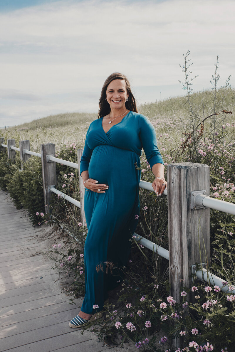 A radiant expectant mother in a flowing blue dress poses along a scenic beach boardwalk, surrounded by wildflowers, during a maternity photography session in Connecticut. Captured by Nicole Mele Photography, embracing the beauty of motherhood in nature.