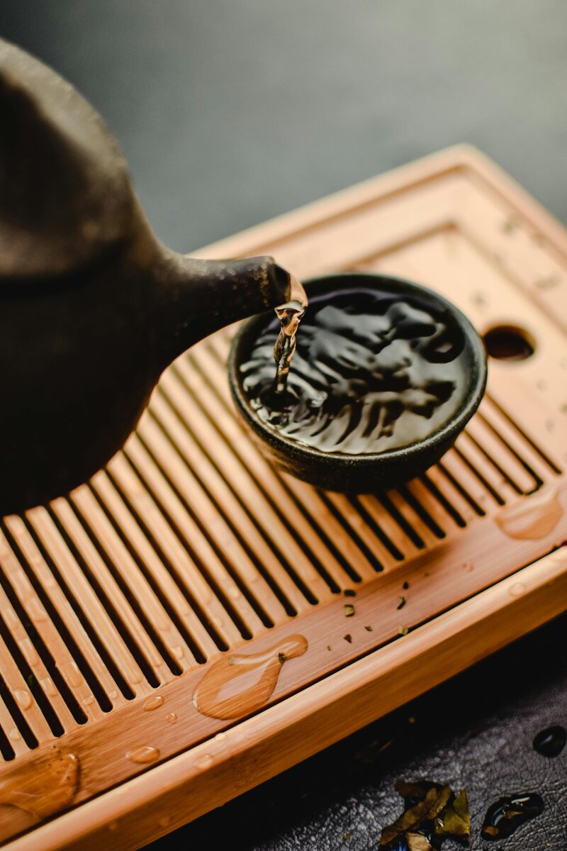 A Chinese black ceramic tea pot pours tea into cup sitting atop a wooden slated board.