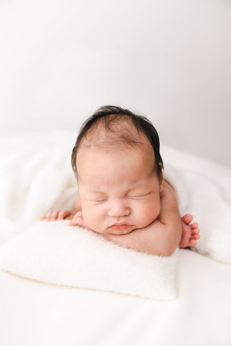 A peaceful newborn baby resting on a soft white blanket with hands gently placed under the chin.