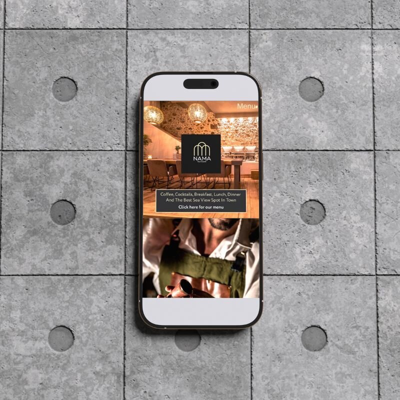 Smartphone on a tiled concrete floor showing a restaurant website with the NAMA logo, a cosy dining space, and a man in an apron holding a cocktail tool.