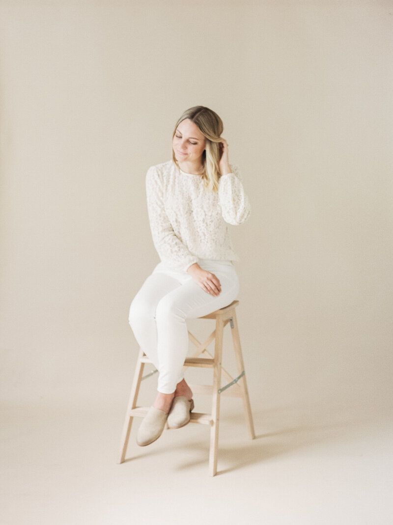 A woman sitting on a wood stool in white clothing.