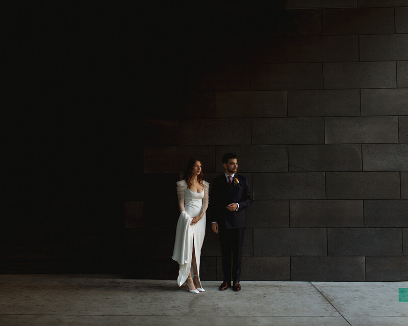 Bride and groom standing against a black wall with moody lighting