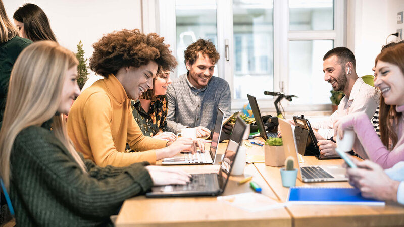 Happy coworkers collaborating on laptops around a shared desk in a modern, creative workspace.
