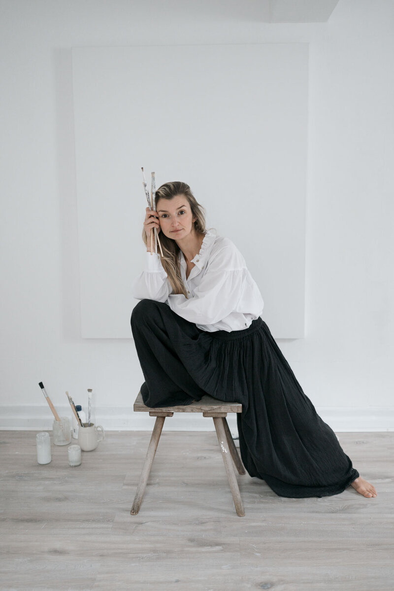 eine Künstlerin in weißer Bluse und schwarzer Flatterhose posiert auf ihrem Hocker mit Pinseln in ihrem Atelier