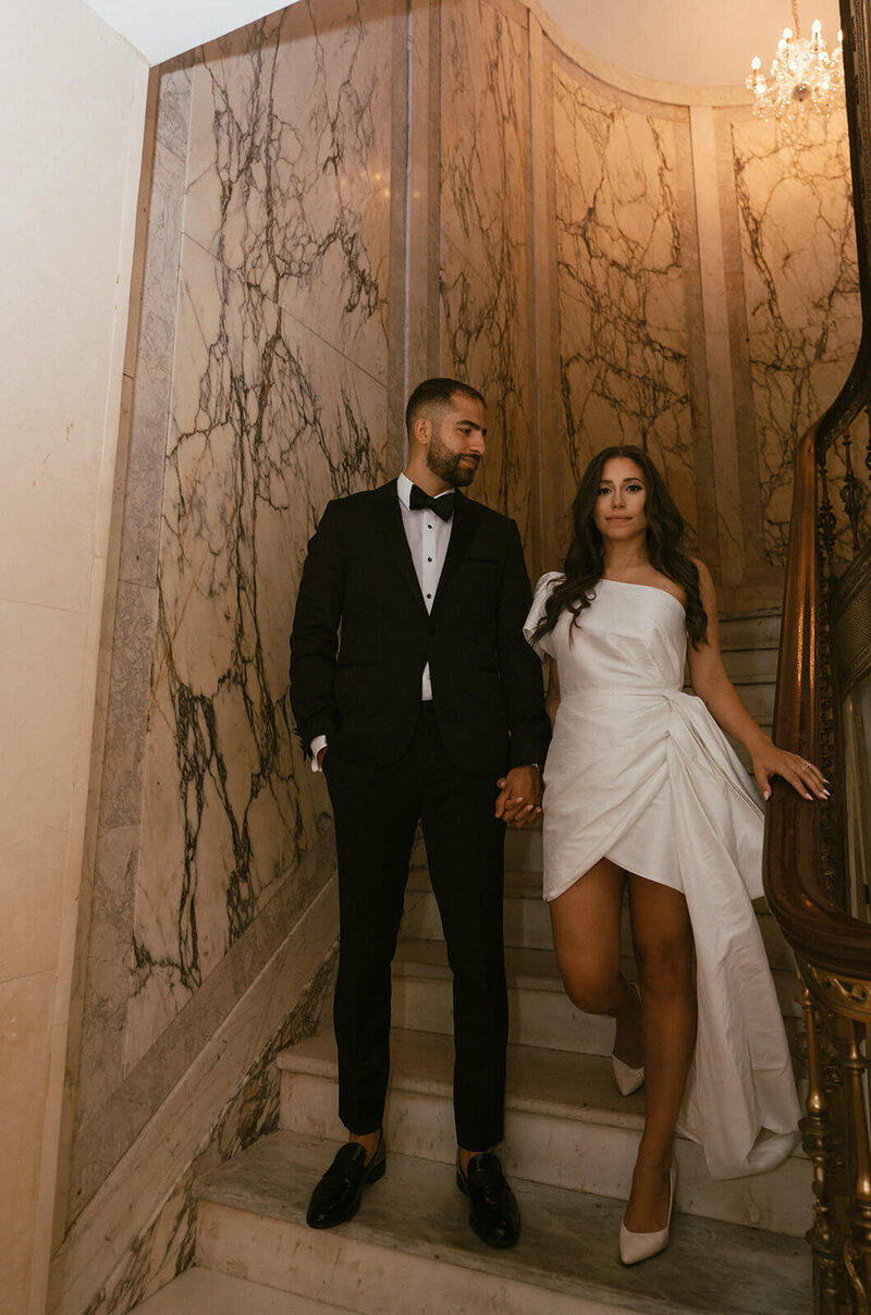 The groom and bride are standing on the staircase, holding hands. The groom is looking at the bride, while the bride is looking at the camera.