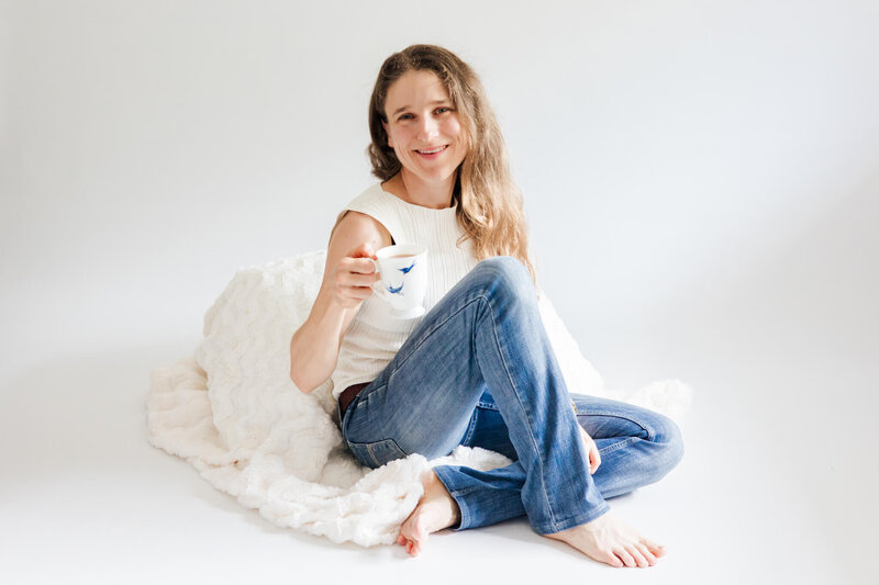 Woman sitting down on a cream blanket, wearing blue jeans and a cream top with her legs crossed. She is holding a white china cup in one hand, smiling.