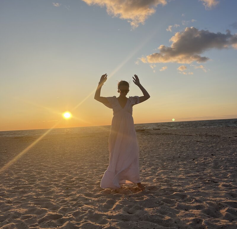 Woman dances in the sand at the beach at sunset with her arms overhead while wearing a flowy, white dress.