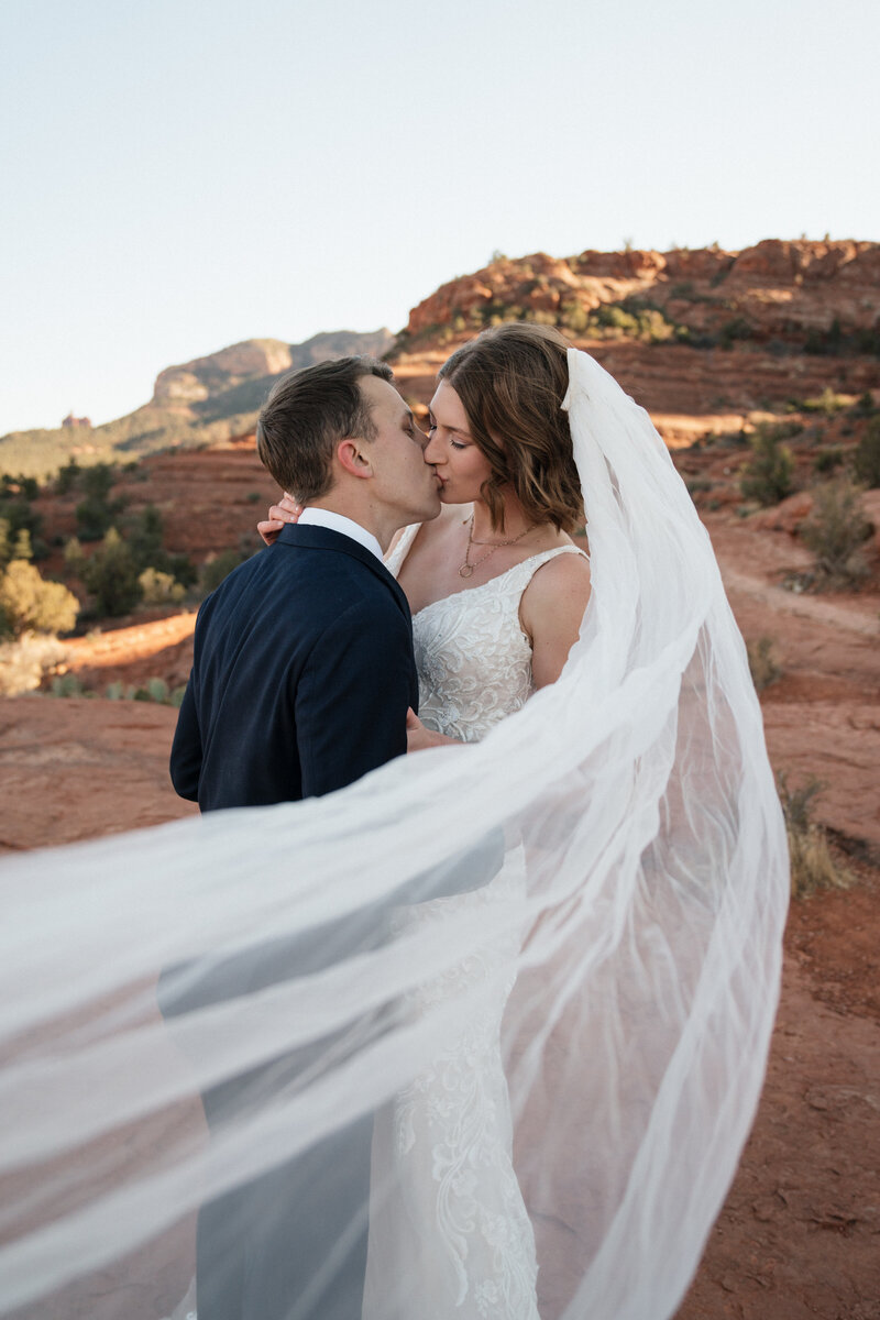 Bride and groom kissing with flowing veil during their Sedona elopement at Cathedral Rock, surrounded by red rock formations and golden light.