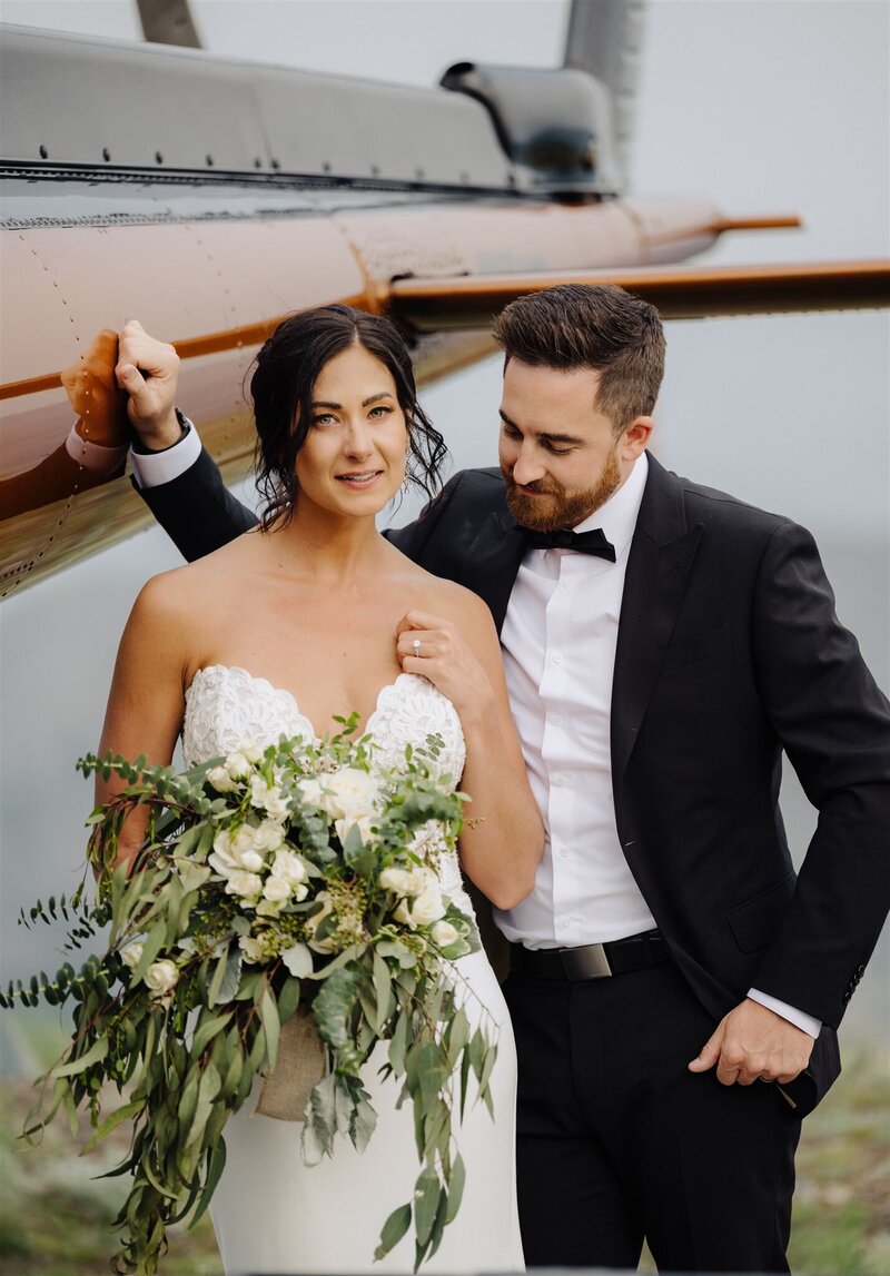 portrait of a married couple after their elopement in Osoyoos - the bride is wearing a white strapless wedding  dress holding a bouquet of white flowers with greenery, and the groom is wearing a black suit. They are standing in front of an orange helicopter