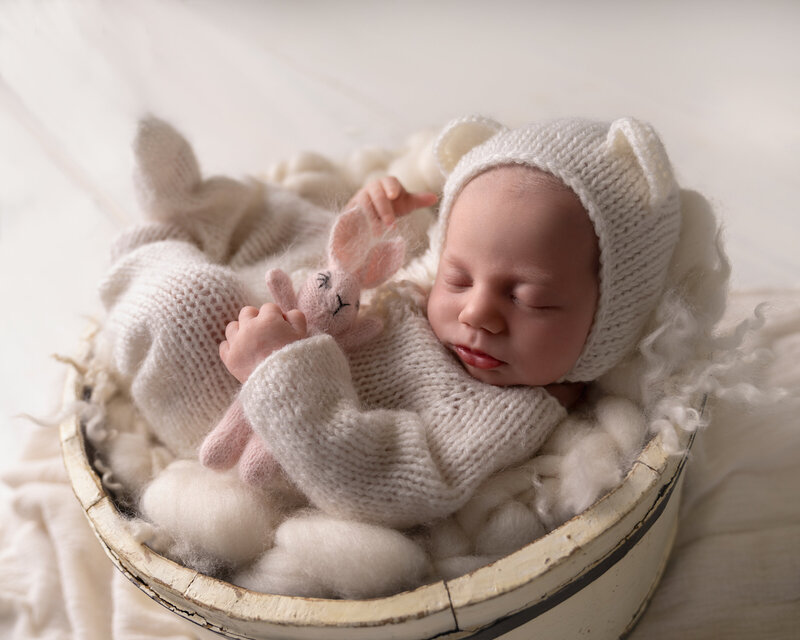 Nyfødt baby i hvit strikkedress og lue med ører, sover i kurv fylt med ull under fotografering nær Oslo.
