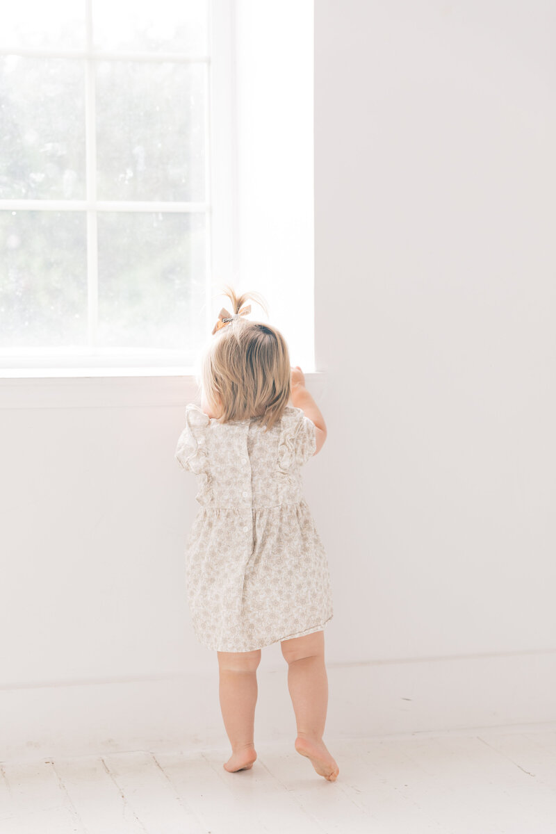 Young girl standing at a window in a soft floral dress during a family session.