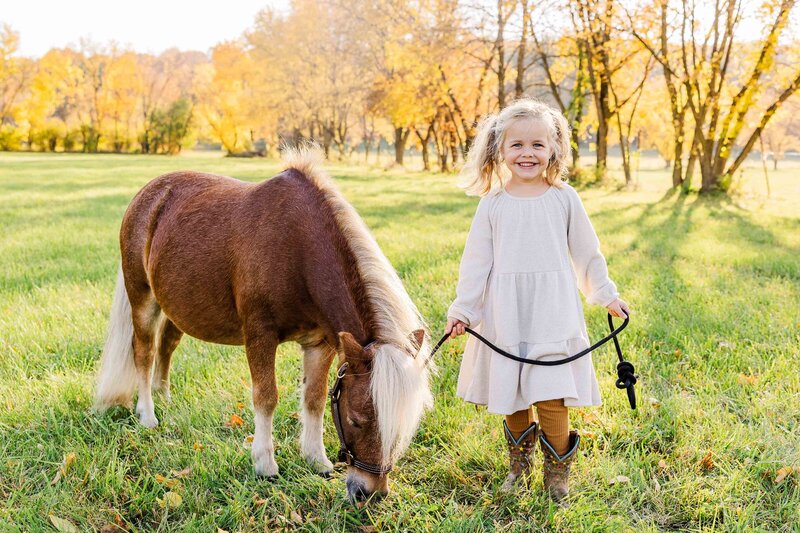 Little girl smiling at camera holding rope connected to a miniature horse in Fall.  Waukesha Family Photographer.