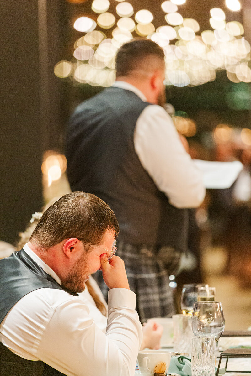 Scottish groom is laughing with hands over his eyes sitting next to his Jewish bride at their sweetheart table while his Scottish best man is giving a toast  in the ballroom at District Winery in DC 