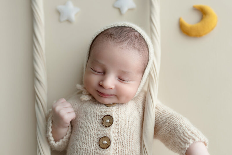Baby boy posed on a swing with celestial moon-and-star props, dressed in a cozy cream knit outfit—dreamy and gentle.