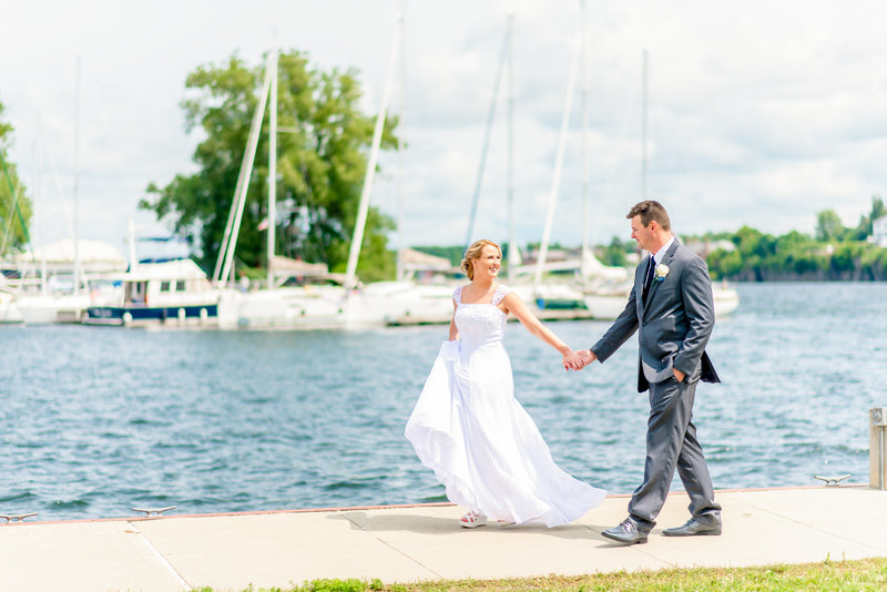 A bride and groom walk hand in hand in front of sailboats