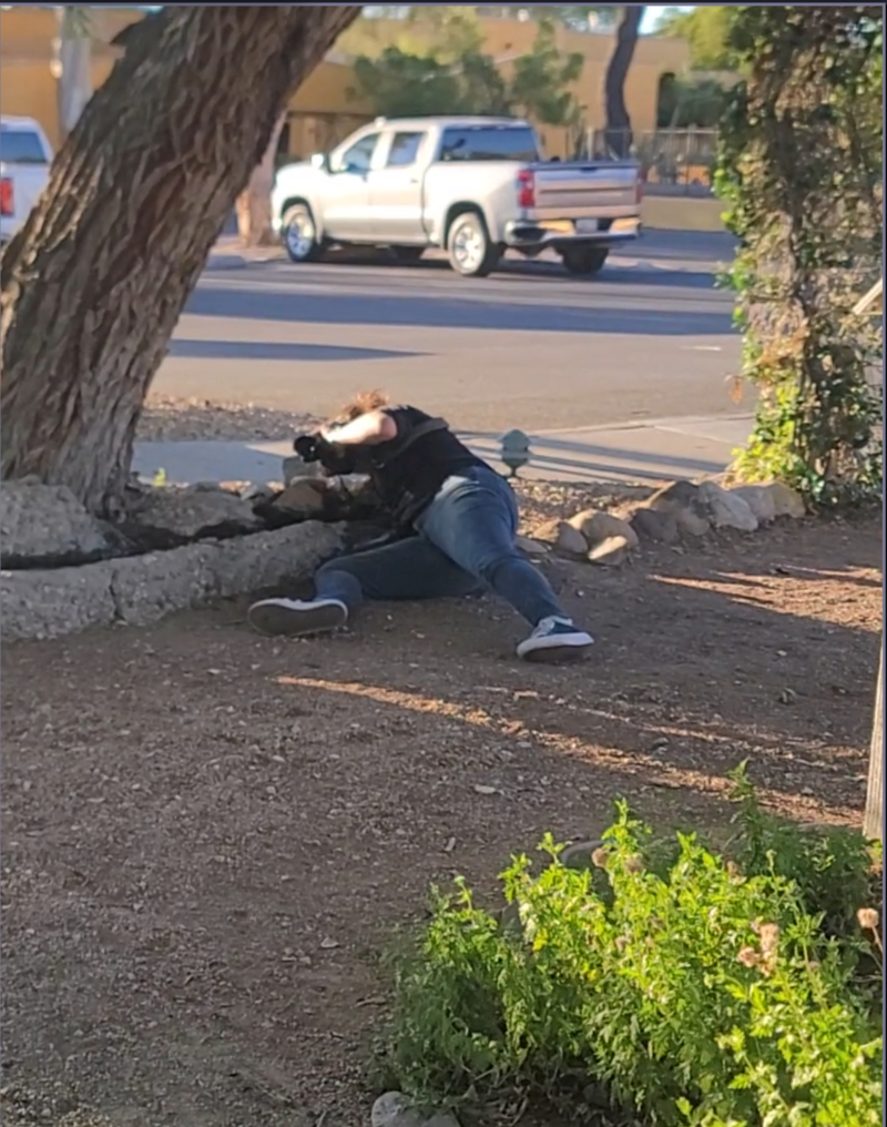 A behind-the-scenes moment of a photographer lying on the ground outdoors to capture a creative shot, with trees and a street scene in the background.