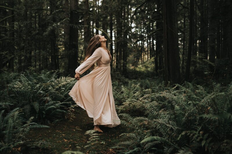 Woman stands in dark, lush forest wearing a cream-colored flowy dress with arms outstretched behind her.