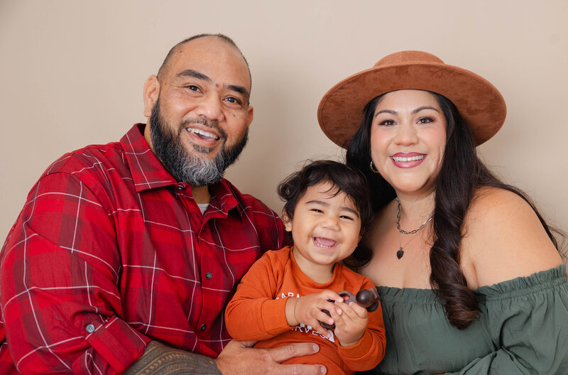 Mom, Dad, and toddler boy posed for family photo with a beige background. 