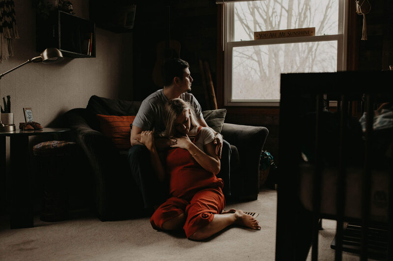 A couple sits together on the floor, the woman resting her head on the man's shoulder as he looks out the window. ​​