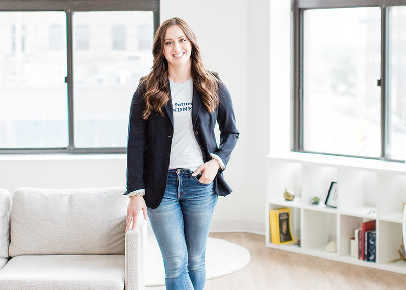 photo of the brand photographer, Laurelyn of My Brand Photographer, standing in a bright studio with one hand in her pocket and one on a gray couch in a casual stance, wearing jeans and a navy blue blazer