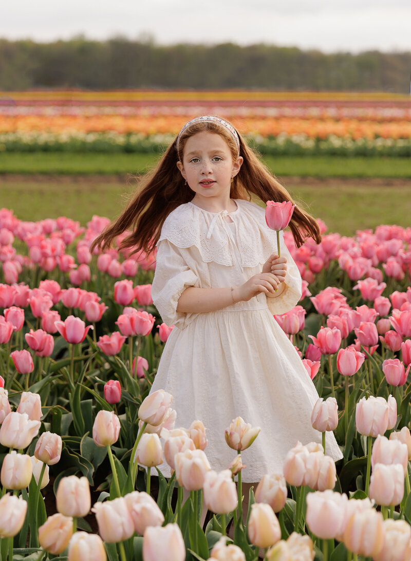 Outdoor photoshoot in NYC. Little girl with long red hair is standing in a field of tulips. She is twirling and holding a flower. Captured by best Brooklyn, NY family photographer Chaya Bornstein.